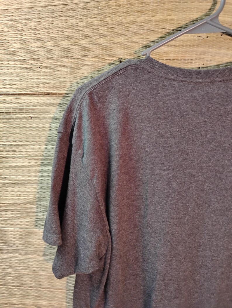 Puede incluir: Una camiseta gris jaspeado cuelga de una percha de pl&aacute;stico gris. La camiseta de manga corta est&aacute; hecha de un material suave. Se ven el cuello y las costuras de los hombros de la camiseta. El fondo es una superficie tejida de color marr&oacute;n claro.