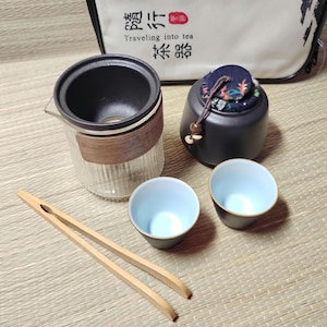 Könnte beinhalten: Ein Teeservice mit einer Glasteekanne, zwei kleinen Tassen mit blauem Rand, einem schwarzen Keramikbehälter und Holzpinzetten. Die Teekanne hat ein Holzband. Eine Tasche mit chinesischen Schriftzeichen befindet sich im Hintergrund. Das Set steht auf einer gewebten Matte.