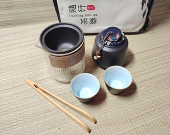 Puede incluir: Un juego de té con una tetera de vidrio, dos tazas pequeñas con borde azul, un recipiente de cerámica negro y pinzas de madera. La tetera tiene una banda de madera. Una bolsa con caracteres chinos está en el fondo. El juego está sobre una estera tejida.