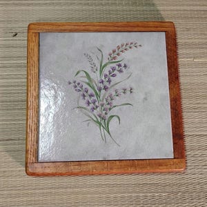 Pode incluir: Tábua de madeira quadrada com um design floral. O azulejo central apresenta uma pintura em aquarela de flores roxas e folhas verdes. A tábua é emoldurada por uma borda de madeira castanho-claro.