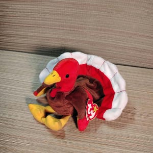 Puede incluir: Un juguete de peluche de pavo con cabeza roja, pico y patas amarillas y cuerpo marrón. El pavo tiene una cola blanca, roja y marrón. Una etiqueta roja está adherida al cuerpo del pavo.