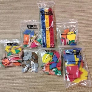 May include: A collection of six clear plastic bags containing various educational math manipulatives, including colorful plastic pattern blocks, attribute blocks, interlocking cubes, and a small pile of assorted metal coins for classroom learning.