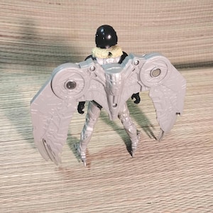 Pu&ograve; includere: Action figure grigia di un personaggio con grandi ali dettagliate. La figura ha un casco nero, un colletto beige e un corpo argentato e grigio. Le ali hanno un design meccanico con dettagli circolari.