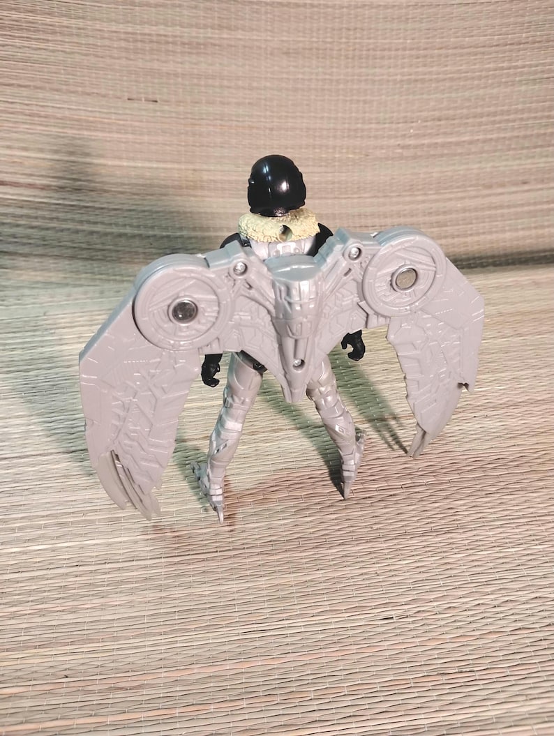Pu&ograve; includere: Action figure grigia di un personaggio con grandi ali dettagliate. La figura ha un casco nero, un colletto beige e un corpo argentato e grigio. Le ali hanno un design meccanico con dettagli circolari.