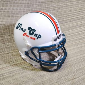 Puede incluir: Un mini casco de fútbol americano blanco con una máscara azul y una franja roja y naranja. El casco tiene el texto "Fins Cup Pro-am" en una fuente retro. Un artículo de colección.