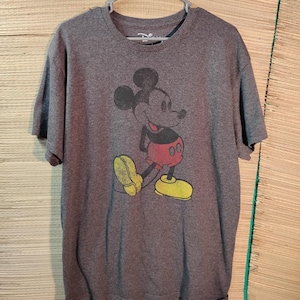 Puede incluir: Una camiseta gris con un gr&aacute;fico de Mickey Mouse de estilo vintage. El personaje de dibujos animados est&aacute; representado en negro, rojo y amarillo. La camiseta est&aacute; colgada de una percha de pl&aacute;stico gris.