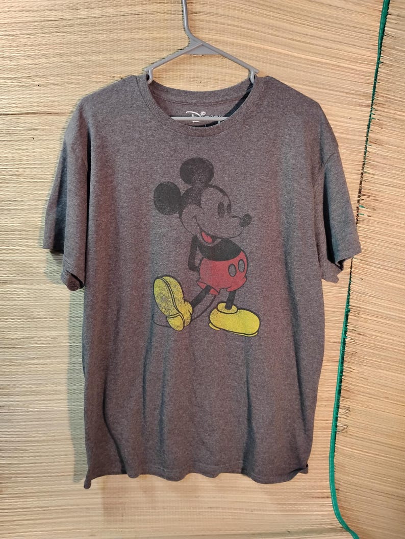 Puede incluir: Una camiseta gris con un gr&aacute;fico de Mickey Mouse de estilo vintage. El personaje de dibujos animados est&aacute; representado en negro, rojo y amarillo. La camiseta est&aacute; colgada de una percha de pl&aacute;stico gris.