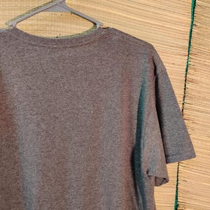 Puede incluir: Una camiseta gris jaspeada de manga corta colgada de una percha de pl&aacute;stico gris. La camiseta est&aacute; hecha de una tela suave y texturizada. El fondo es una estera tejida.