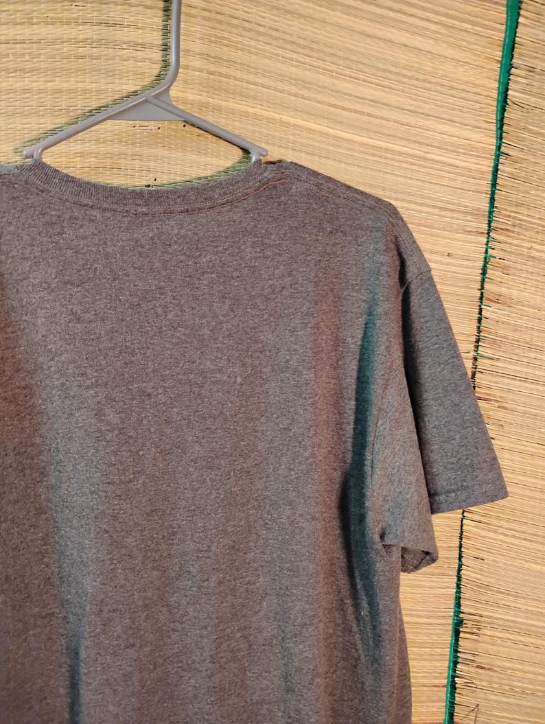 Puede incluir: Una camiseta gris jaspeada de manga corta colgada de una percha de pl&aacute;stico gris. La camiseta est&aacute; hecha de una tela suave y texturizada. El fondo es una estera tejida.