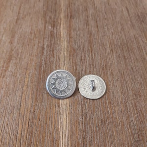 May include: Two silver buttons with a decorative design. One button has a sun-like design with a textured center. The other button has a simple design with a loop on the back.