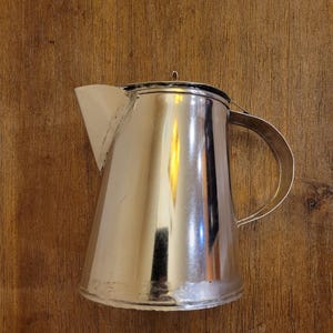 May include: A shiny, silver-colored metal pitcher with a handle and a spout. The pitcher has a conical shape and a lid. The background is a wooden surface.