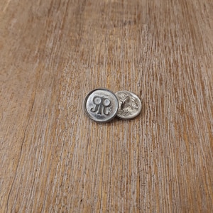 May include: Two silver-toned buttons with a raised "R" design. The buttons are on a wooden surface.