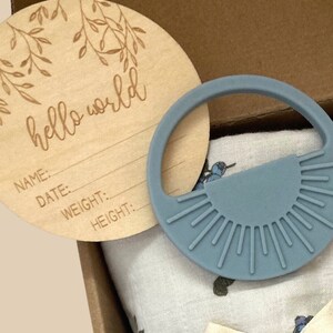 May include: A baby gift set with a wooden engraved round plaque that says "hello world" with lines for name, date, weight, and height. The set also includes a blue silicone teether and a white muslin swaddle with a blue floral pattern.