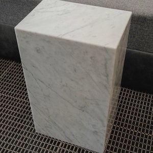 May include: A white marble cube-shaped side table with a smooth, polished surface. The table is sitting on a black and brown patterned rug.