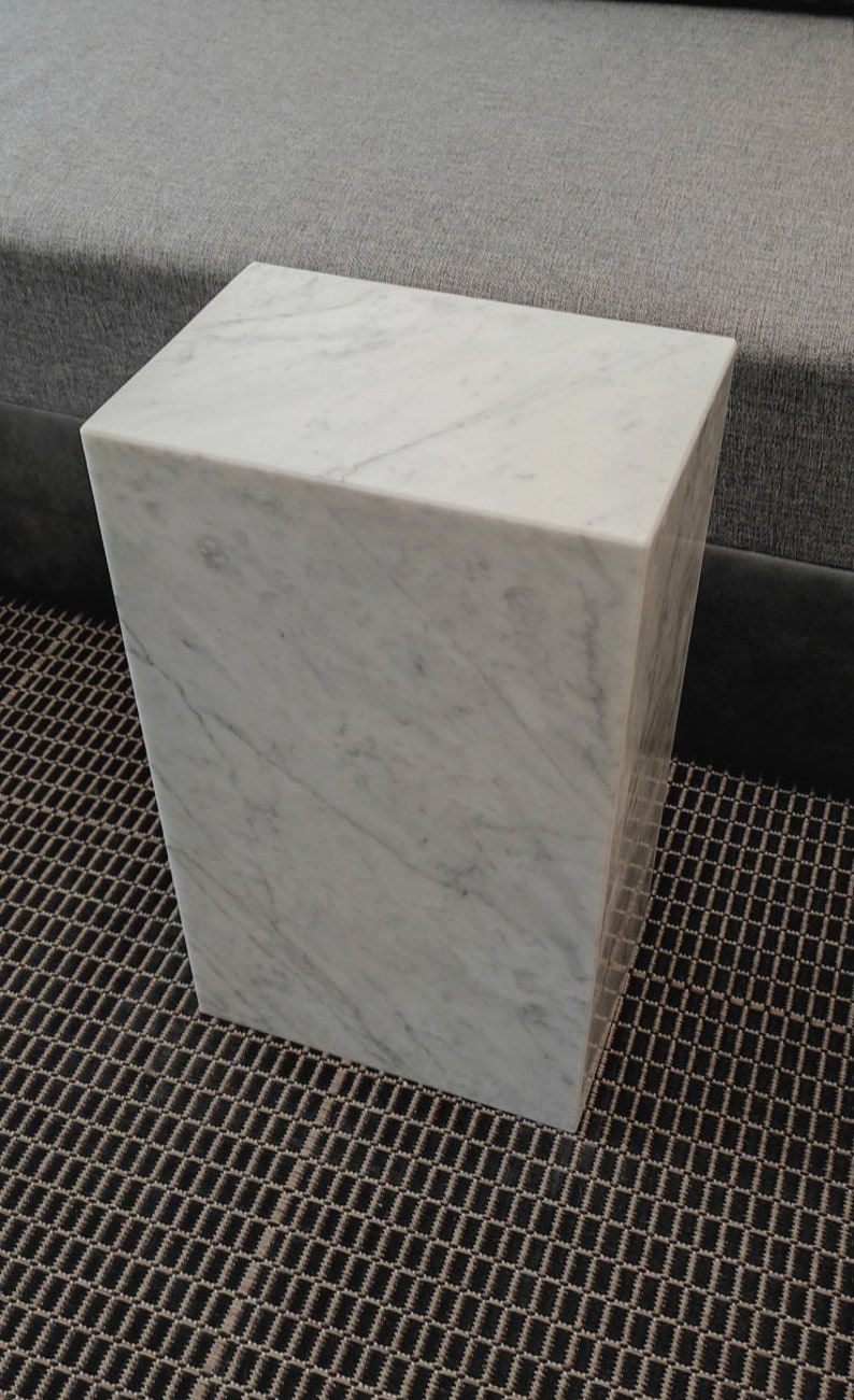 May include: A white marble cube-shaped side table with a smooth, polished surface. The table is sitting on a black and brown patterned rug.