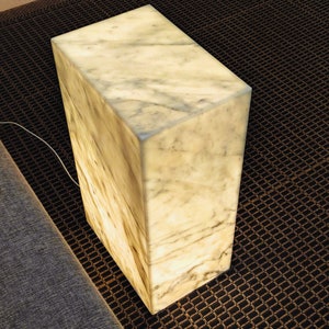 May include: A white marble cube lamp with a warm, glowing light. The lamp is sitting on a black and brown patterned rug.