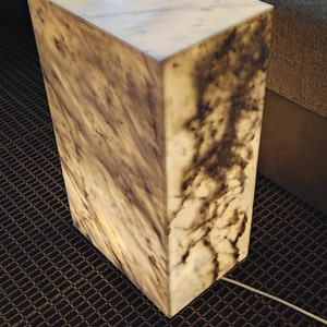 May include: A white marble cube lamp with a light brown marbling pattern. The lamp is sitting on a dark brown carpet with a diamond pattern.