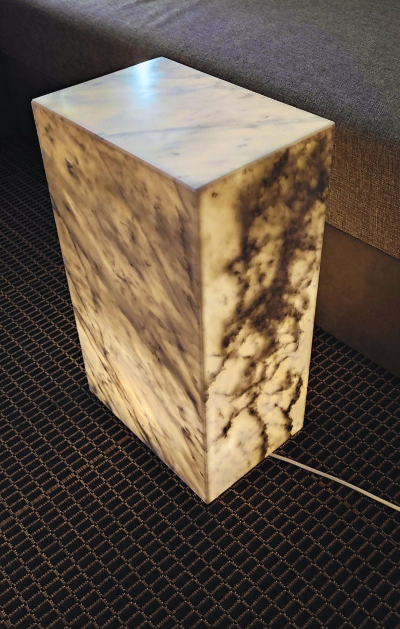 May include: A white marble cube lamp with a light brown marbling pattern. The lamp is sitting on a dark brown carpet with a diamond pattern.