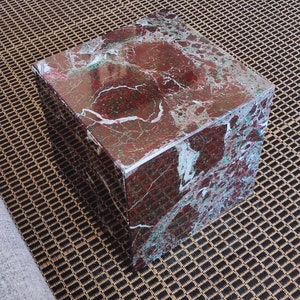 May include: A square, red and green marble cube-shaped stool. The stool is sitting on a brown and black patterned rug.