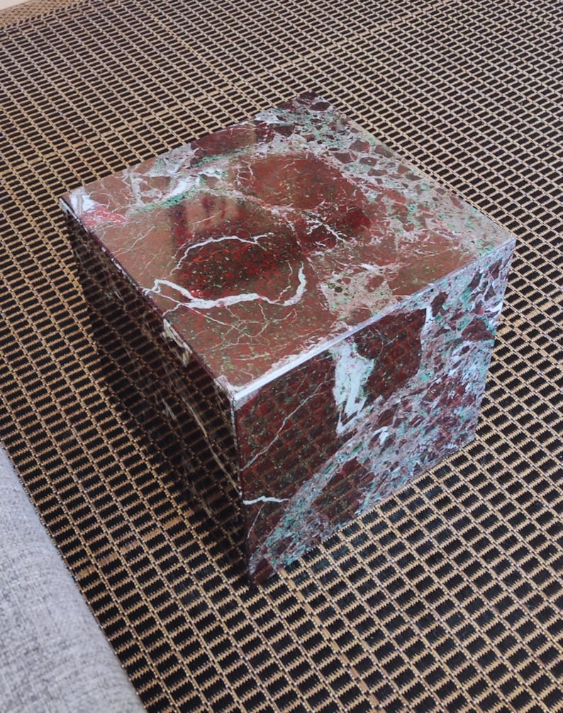 May include: A square, red and green marble cube-shaped stool. The stool is sitting on a brown and black patterned rug.