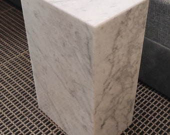 Könnte beinhalten: Ein weißer Marmor-Beistelltisch mit quadratischer Platte und rechteckigem Sockel. Der Tisch steht auf einem braunen Teppich mit Muster.