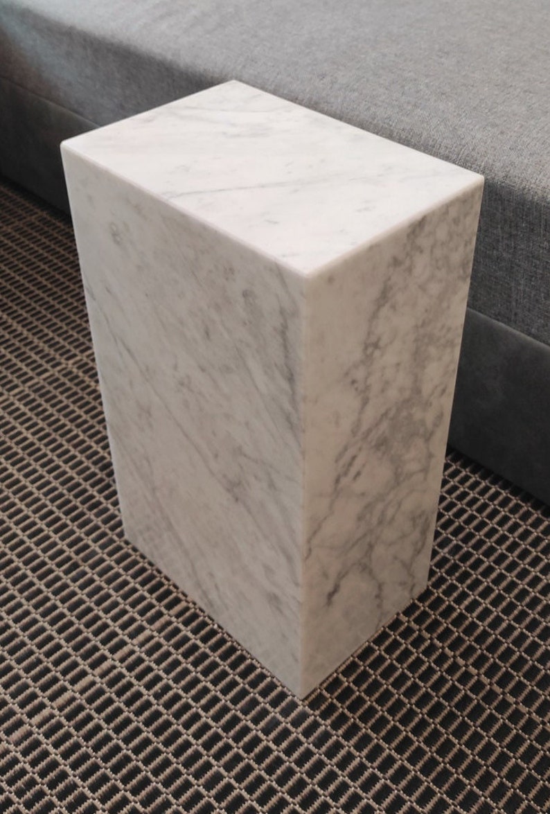May include: A white marble side table with a square top and a rectangular base. The table is sitting on a brown patterned rug.