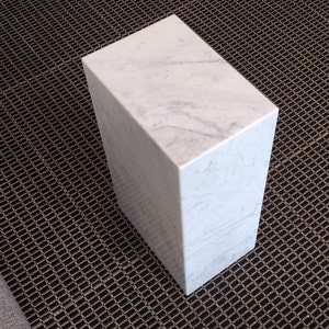 May include: A white marble cube-shaped side table with a smooth, polished surface. The table is sitting on a black metal grate.