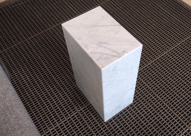May include: A white marble cube-shaped side table with a smooth, polished surface. The table is sitting on a black metal grate.