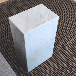 May include: A white marble cube pedestal with a smooth, polished surface. The pedestal is sitting on a brown woven rug.