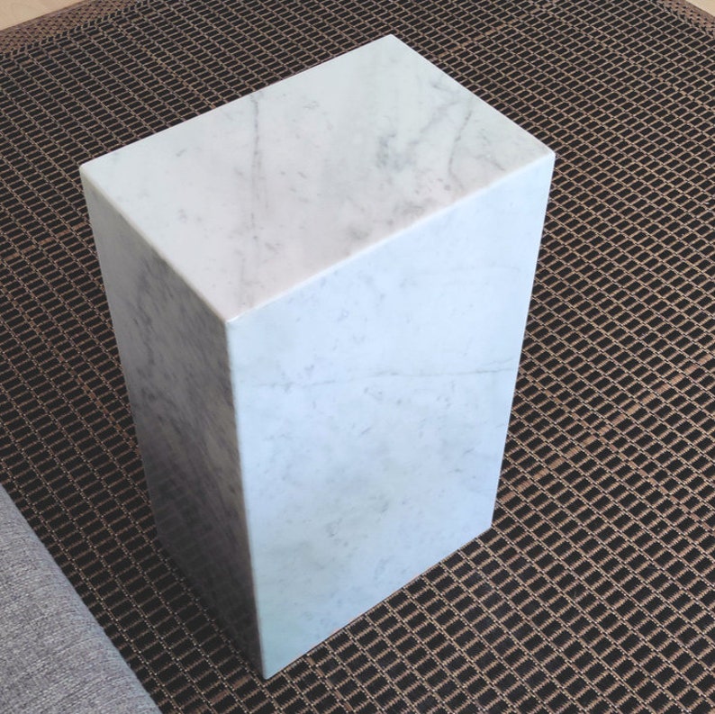 May include: A white marble cube pedestal with a smooth, polished surface. The pedestal is sitting on a brown woven rug.