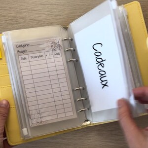 May include: Yellow binder-style budget planner with clear plastic pockets. One pocket contains a lined sheet with prompts for category, budget, date, description, and sale. Another pocket has the word "Cadeaux" written on a white card.