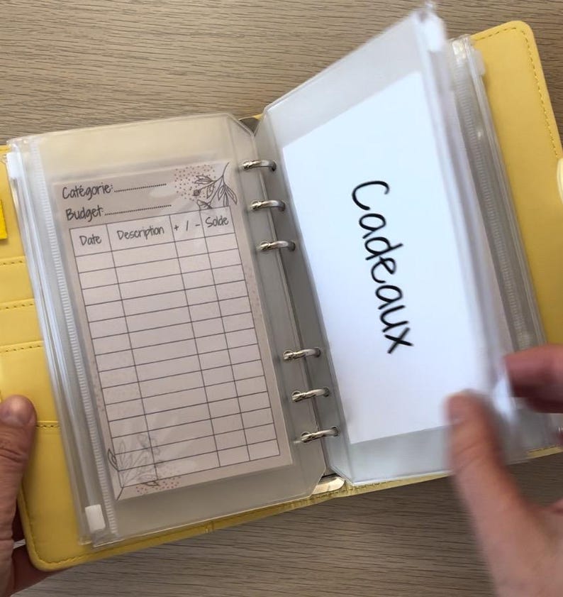 May include: Yellow binder-style budget planner with clear plastic pockets. One pocket contains a lined sheet with prompts for category, budget, date, description, and sale. Another pocket has the word "Cadeaux" written on a white card.
