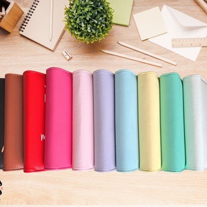 May include: A collection of colorful, leather-bound journals in various shades, including black, brown, red, pink, lavender, blue, yellow, green, and white. The journals are arranged on a wooden surface, with stationery and a small plant in the background.
