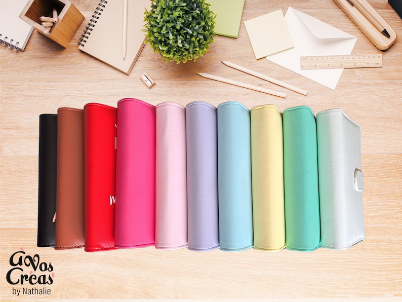 May include: A collection of colorful, leather-bound journals in various shades, including black, brown, red, pink, lavender, blue, yellow, green, and white. The journals are arranged on a wooden surface, with stationery and a small plant in the background.
