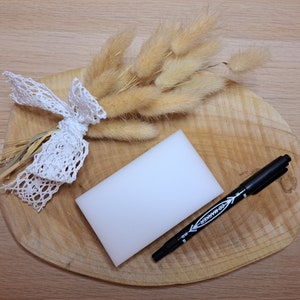 May include: A still life arrangement featuring a wooden cutting board with a white rectangular sponge, a black marker, and a bundle of dried, beige bunny tail grass tied with a white lace ribbon.