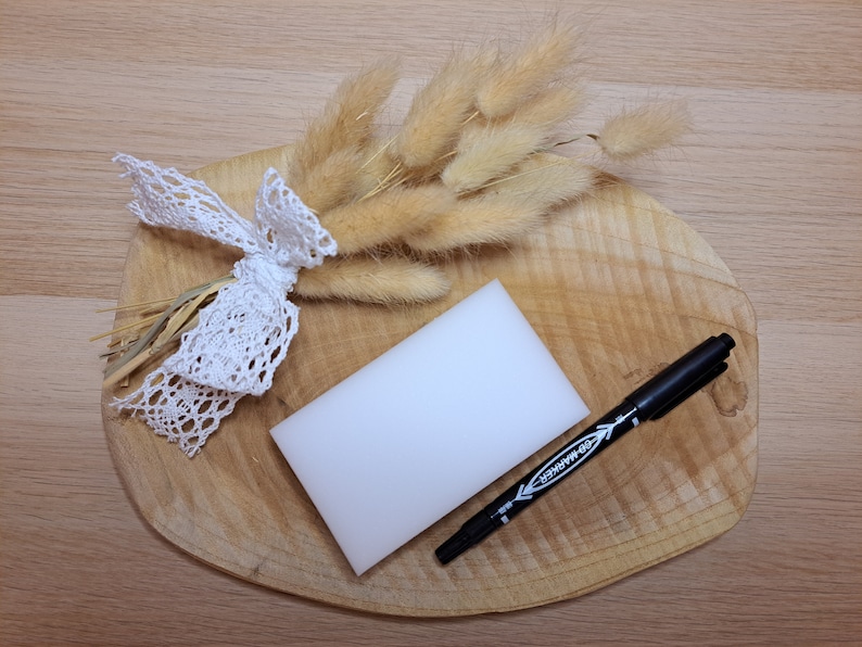 May include: A still life arrangement featuring a wooden cutting board with a white rectangular sponge, a black marker, and a bundle of dried, beige bunny tail grass tied with a white lace ribbon.