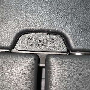 May include: Close-up of a textured black car interior component. The text "GP86" is embossed on a small, rectangular section. The surrounding surfaces are also black and have a leather-like appearance.