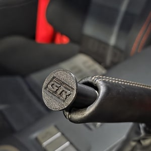 Puede incluir: Primer plano de un pomo de palanca de cambios plateado con las letras "GR" en relieve. El pomo está unido a un mango de cuero negro. El fondo muestra el interior de un coche con asientos negros y detalles en rojo.