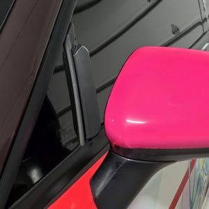 May include: A close-up of a car's side mirror, painted a vibrant pink. The mirror is mounted on a black base, with a red and white section of the car's body visible below. The car's window frame is black.