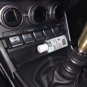 May include: A black car dashboard with climate control knobs and buttons. A small bottle of hand sanitizer with a white cap rests in a storage compartment. The gear shift is visible on the right.