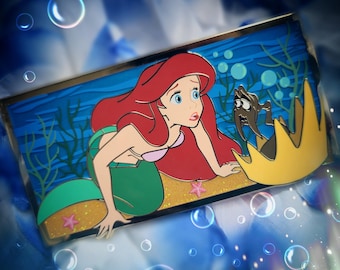 May include: A rectangular enamel pin featuring a cartoon mermaid with long red hair and a green tail, positioned on a sandy ocean floor with a small brown creature and a yellow crown. The background depicts blue water with green seaweed.