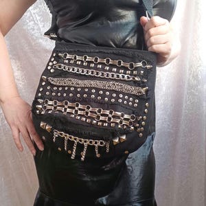 Può includere: Una borsa a tracolla nera con una superficie in tessuto usurato, decorata con diverse file orizzontali di catene metalliche color argento, borchie e finiture decorative. La borsa include una spessa tracolla nera e bordi sfrangiati.