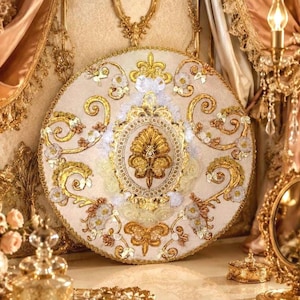 Puede incluir: Un tapiz circular de color crema con intrincados bordados dorados, aplicaciones florales blancas y un motivo central de filigrana dorada. El diseño incluye patrones de volutas doradas, pequeñas rosetas blancas y un borde decorativo.