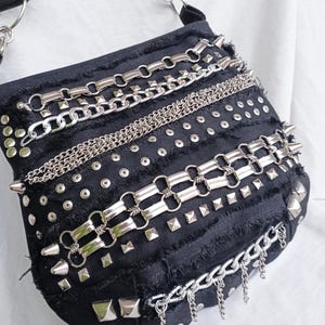 May include: A black crossbody bag featuring frayed edges and a variety of silver-toned metal embellishments, including multiple horizontal chains, rows of circular studs, square pyramid studs, and pointed spikes attached to the front panel.