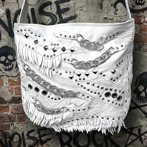 May include: White shoulder bag with a punk rock aesthetic. The bag features silver-tone chain accents, studded details, and fringe trim. The bag has a long strap and is set against a graffiti background.