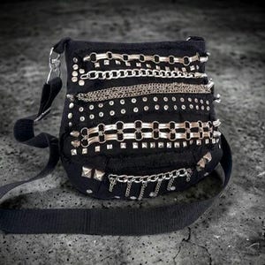 Może przedstawiać: Czarna torba na ramię z srebrnymi łańcuchami, ćwiekami i nitami. Torba ma długi, regulowany pasek i zapięcie na klapę. Torba jest wykonana z teksturowanej tkaniny i ma estetykę grunge.