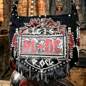 Puede incluir: Bolso de hombro negro con el logo de AC/DC en rojo y blanco. El bolso presenta las palabras "LET'S ROCK" y está decorado con tachuelas metálicas, cadenas y flecos. El bolso tiene una correa negra.