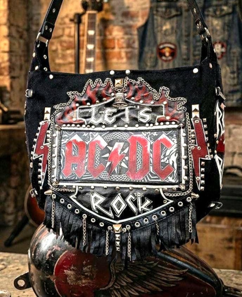 Puede incluir: Bolso de hombro negro con el logo de AC/DC en rojo y blanco. El bolso presenta las palabras "LET'S ROCK" y est&aacute; decorado con tachuelas met&aacute;licas, cadenas y flecos. El bolso tiene una correa negra.