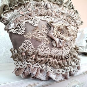 Puede incluir: Bolso decorativo hecho a mano en tonos topo y marrón. Presenta encaje intrincado, volantes y una gran flor de tela. Delicados acentos rosas complementan el diseño, creando una estética vintage y romántica.