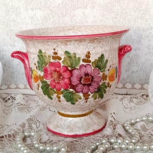 May include: A ceramic planter with a speckled beige finish, featuring two bright pink side handles and a hand-painted floral motif in shades of red, purple, yellow, and green. The vessel has a wide rim, a pedestal base, and a thin gold band.